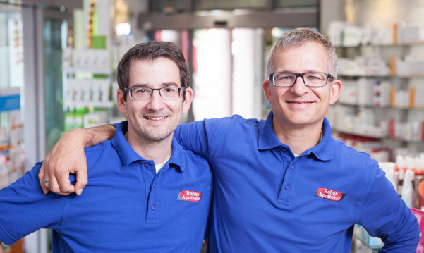 Apotheker Christian Weber und sein Bruder in blauen Arbeitspolohemden lächeln in der hellen und modernen Rohan Apotheke.
