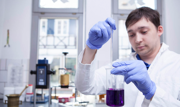 Ein Mitarbeiter der Rohan Apotheke führt eine chemische Analyse in einem Labor durch, indem er eine Pipette verwendet, um eine lila Flüssigkeit in ein Becherglas zu geben, mit Laboreinrichtung im Hintergrund.